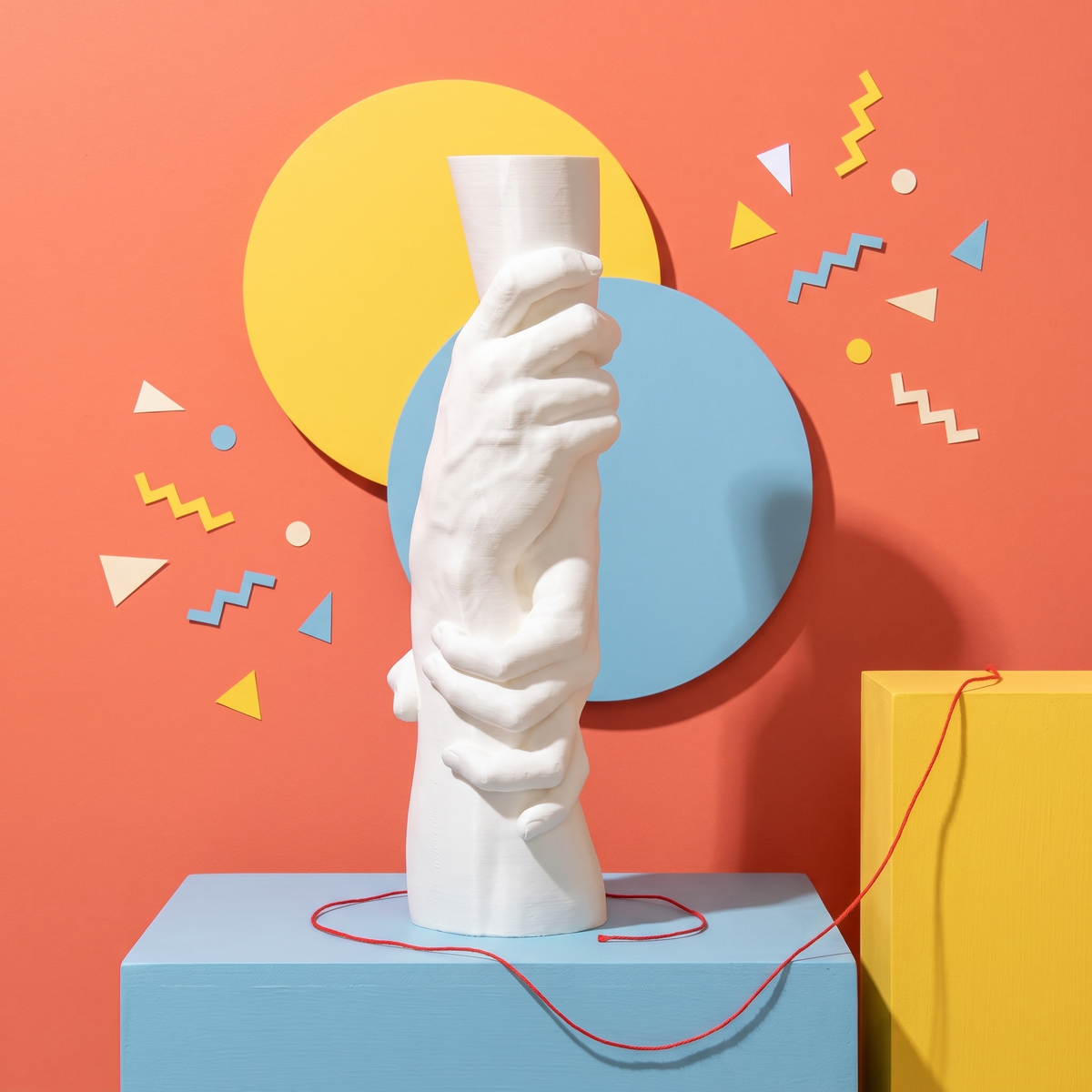 Legame bianco, scultura stampata in 3D di due mani che si stringono il polso, set pop con dischi giallo e azzurro, filo rosso e coriandoli su parete corallo